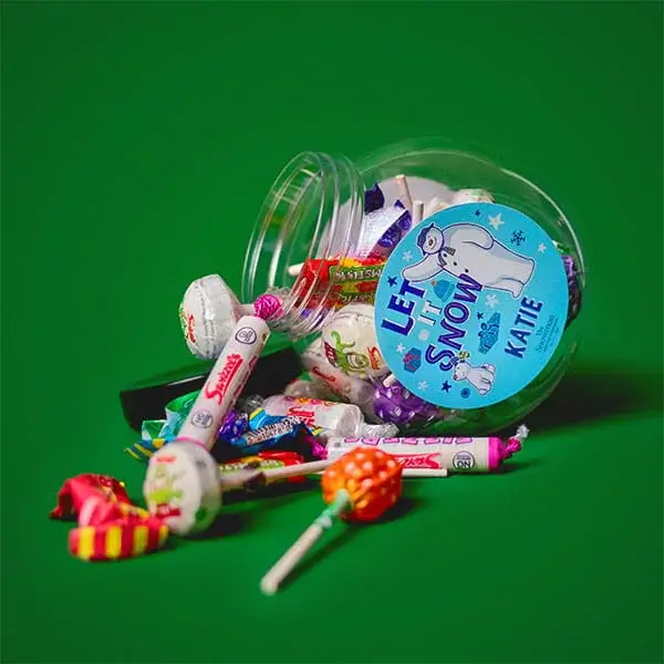 Personalised Christmas sweet jar tipped over with assorted chocolates, lollipops, and festive treats spilling onto a green background.